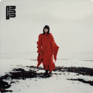 a snowy landscape with a few low black rocks (lava) exposed. A woman with dark hair in a long red dress stands in the middle of the scene