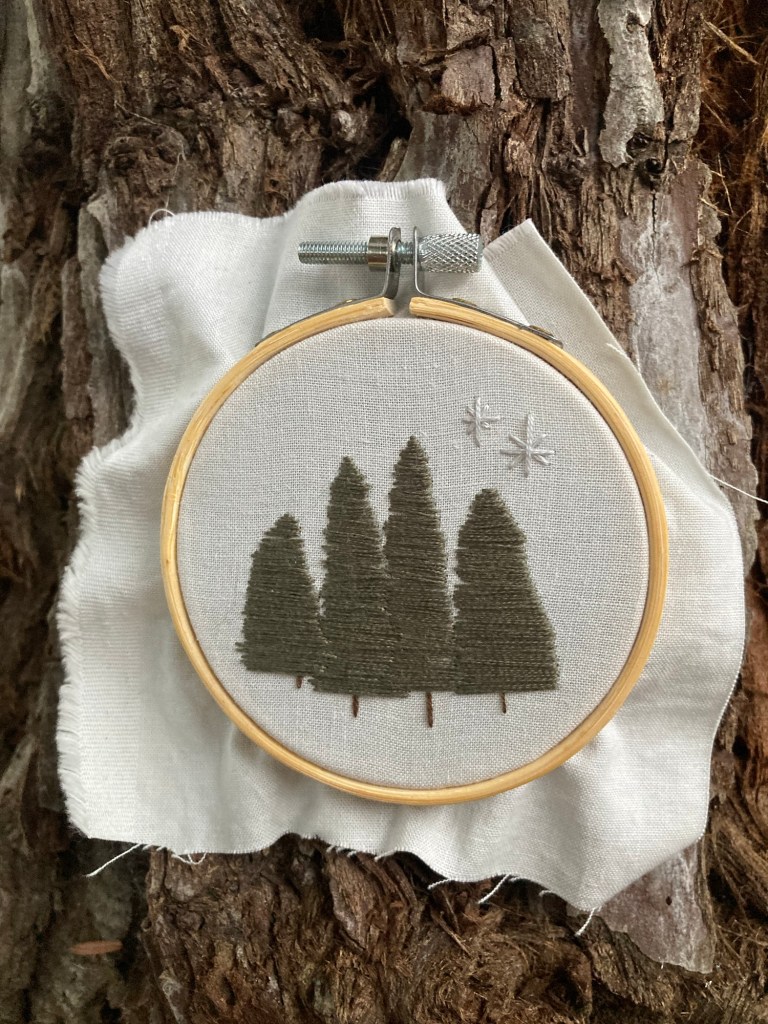 a small embroidery hoop leaned against a tree trunk. the fabric in the hoop is off-white and has 4 embroidered evergreen trees and two stars above them